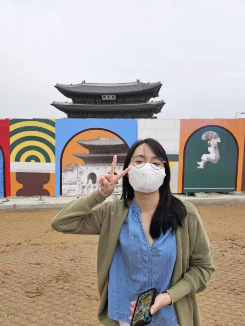 A person with long hair wearing glasses, a blue shirt, and a green sweater, posing for the photo in front of a mural wall with a Chinese building in the background
